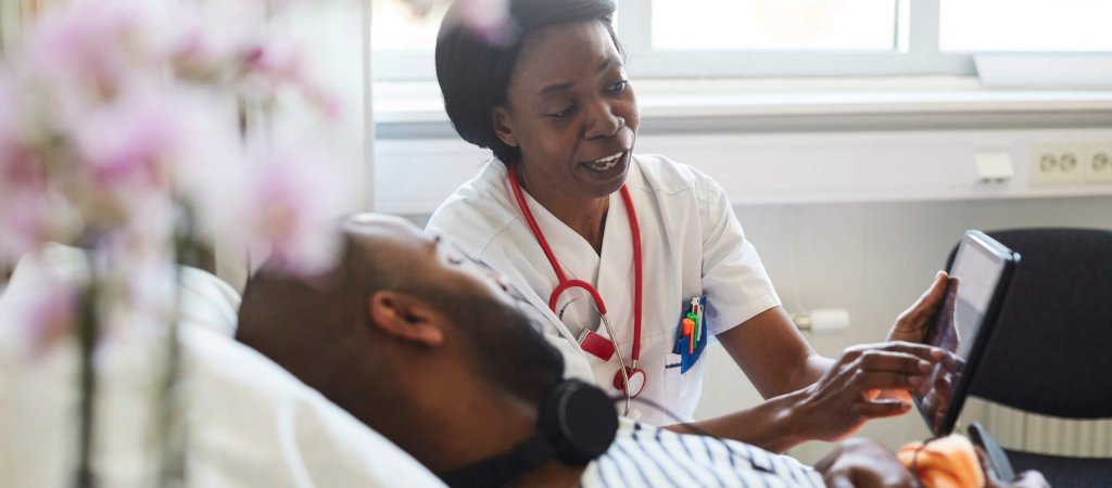 Mature practitioner showing digital tablet to male patient during routine visit in hospital Mature practitioner showing digital tablet to male patient during routine visit in hospital