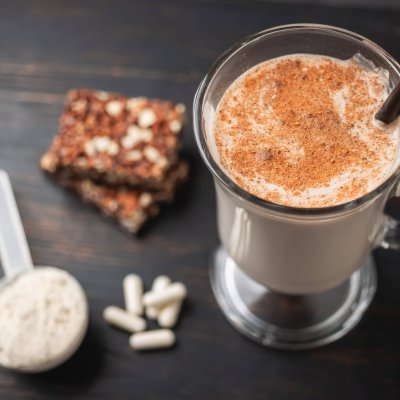 Glass of protein milkshake drink with straw and scoop of whey protein powder, white capsules of amino acids, vitamins, protein bar on a dark wooden board, bodybuilding food supplements