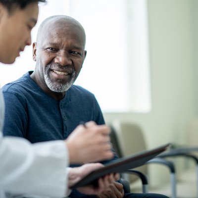A senior gentleman of African decent sits in a waiting room beside his doctor as they review test results on a tablet together.
